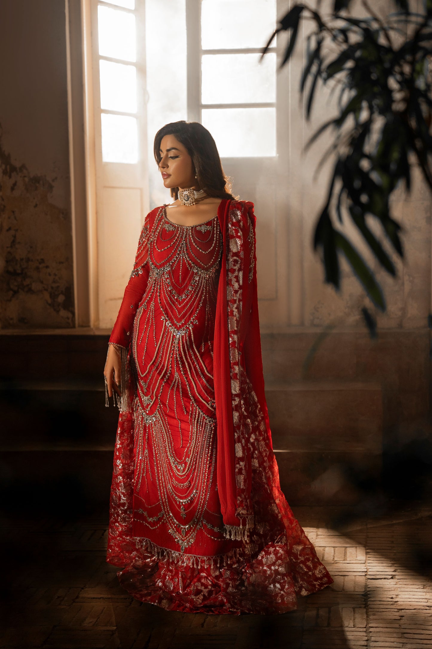 Woman in a red traditional outfit with intricate patterns standing in a softly lit room.