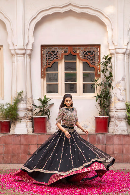 Woman in traditional black and gold outfit standing in front of an architectural building with plants.